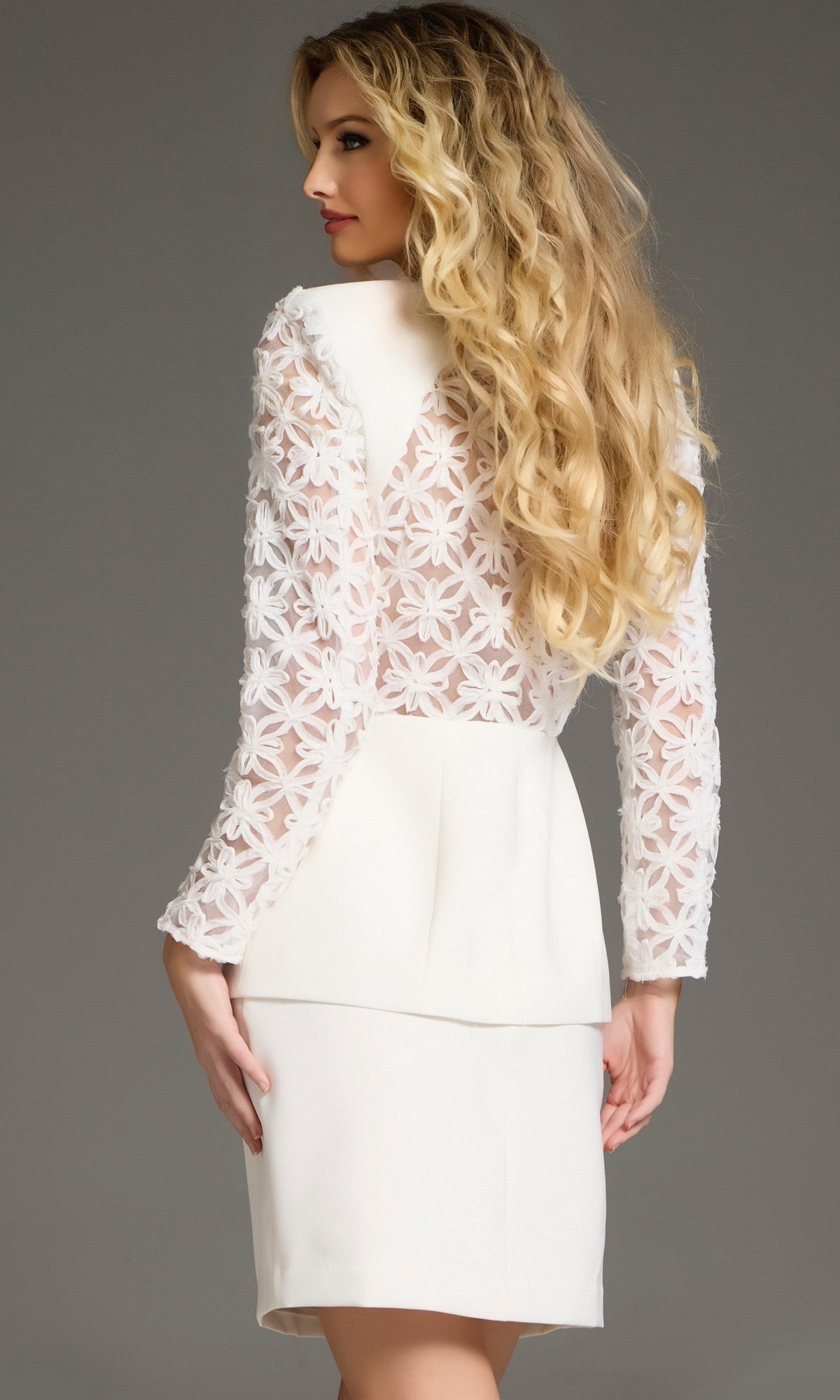 A woman with long blonde hair is wearing the Short Interview Dress: Jovani 39075—a white blazer dress with floral lace back and sleeves—standing against a plain gray background, facing away and looking over her shoulder.